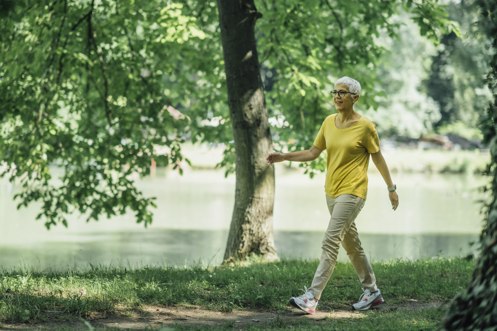 Older woman with grey hair, walking by a lake with trees