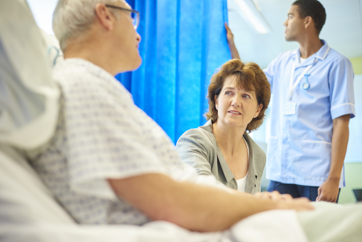 A man sitting in a hospital bed in a hospital bay. He is being consolidated by his wife whilst a male nurse speaks to him.