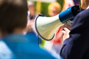 Man holding a megaphone on picket line while speaking to a crowd.