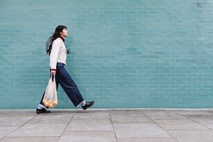 A woman walking with large strides infront of a large blue brick wall carrying a bag of shopping