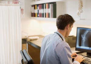 A male doctor is sitting at a desk on the computer in a doctor's surgery